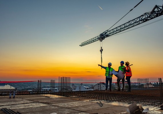Three Multi-Ethnic construction workers in uniform standing at construction site with crane in background, discussing building plans while holding blueprint at sunset under the tower crane.